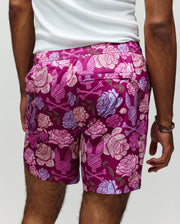 A person stands facing away, wearing Psycho Bunny MENS ROWEN SWIM TRUNK - MB1000023: quick-dry, magenta floral swimwear with bold pink and blue patterns, paired with a white tank top.