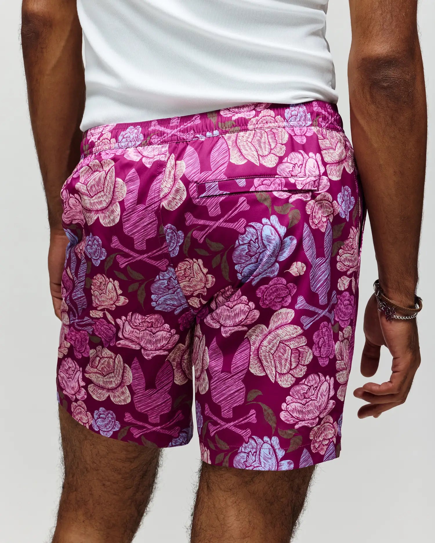 A person stands facing away, wearing Psycho Bunny MENS ROWEN SWIM TRUNK - MB1000023: quick-dry, magenta floral swimwear with bold pink and blue patterns, paired with a white tank top.