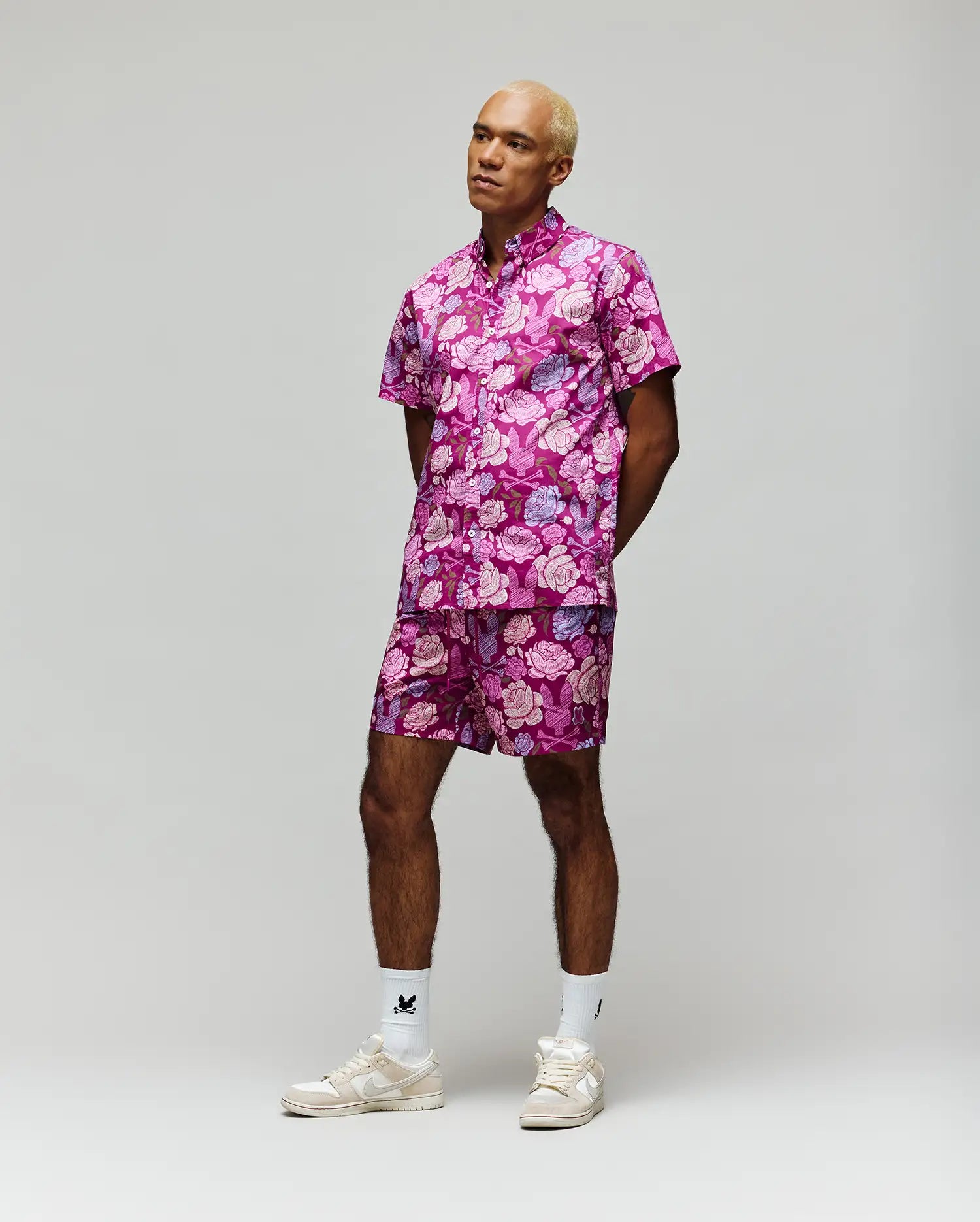 A person with short blond hair wears Psycho Bunny MENS ROWEN SWIM TRUNK - MB1000023, featuring a pink and purple floral print with mesh lining, paired with white crew socks and beige sneakers, standing against a plain light gray background.