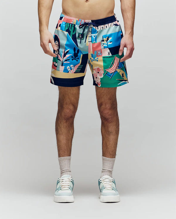 A person wears Psycho Bunny MENS BLAISE SWIM TRUNK - MB1000028 with vibrant tropical patterns and bunny logos, paired with light gray socks and white sneakers, standing against a plain light background.