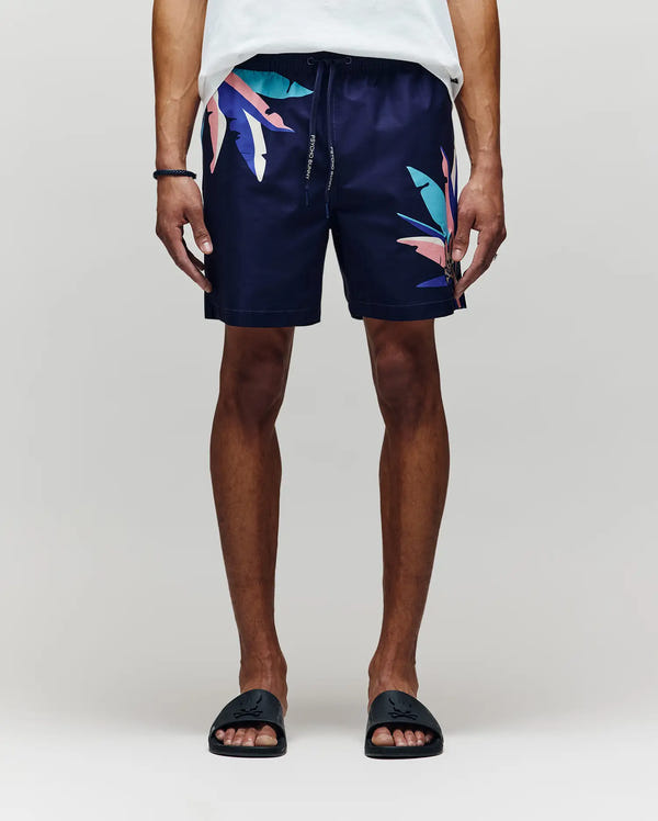 A person stands against a plain background, showing only the lower half, wearing Psycho Bunny MENS SHANE SWIM TRUNK - MB1000029 with colorful abstract leaf designs, paired with a white shirt and black slide sandals for a relaxed, luxurious look.