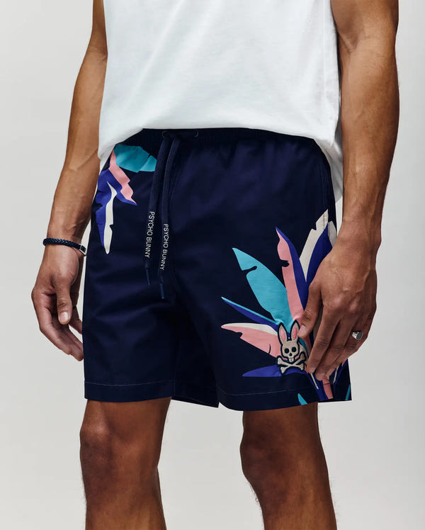 Wearing Psycho Bunny MENS SHANE SWIM TRUNK - MB1000029 in navy with abstract leaf and bunny graphics, paired with a white tee. The drawstring waist adds comfort for luxurious relaxation as he stands with one hand by his side.