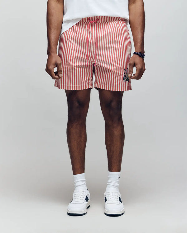 Only the lower body is visible, featuring Psycho Bunny's MENS ZANE SWIM TRUNK - MB1000030 in red and white stripes with an elastic waistband, paired with a white T-shirt, socks, and sneakers against a plain background.