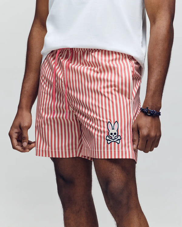 A model wears Psycho Bunny MENS ZANE SWIM TRUNK - MB1000030, featuring red and white vertical stripes and a cartoon bunny & crossbones patch on the left leg, shown from waist to mid-thigh with a white shirt and dark bracelet.