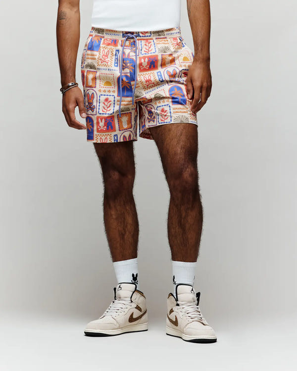 A person stands against a plain light background, showing only their lower body in Psycho Bunny MENS SUNDEW SWIM TRUNK - MB1000040 with colorful abstract graphics, paired with a tucked-in white shirt, white socks, and beige high-top sneakers.