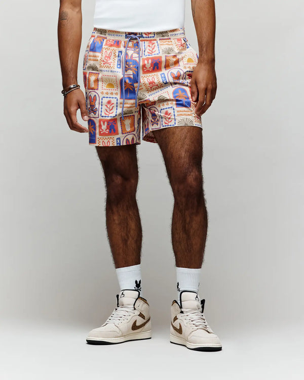 A person stands against a plain light background, showing only their lower body in Psycho Bunny MENS SUNDEW SWIM TRUNK - MB1000040 with colorful abstract graphics, paired with a tucked-in white shirt, white socks, and beige high-top sneakers.