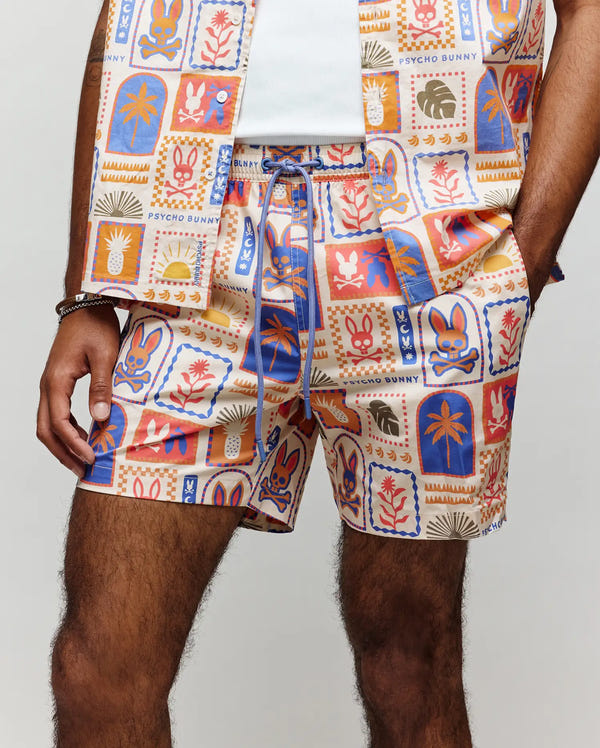A person wears Psycho Bunny's bold MENS SUNDEW SWIM TRUNK (MB1000040) and a matching shirt, both featuring a whimsical multicolored pattern of bunnies, flowers, and abstract shapes with the brand name visible.