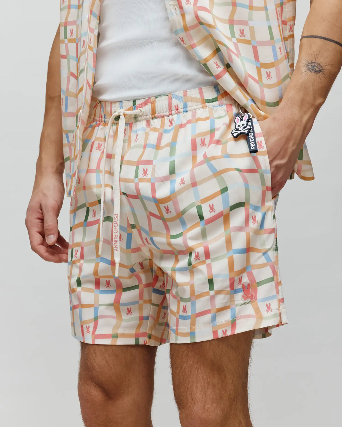 Mens Atlas Swim Trunk in cream, featuring a vibrant multi-color wavy plaid pattern with repeated small bunny skull logos, relaxed fit, smooth quick-dry fabric, drawstring waist with Psycho Bunny text, and an embroidered pink bunny skull on the left leg.