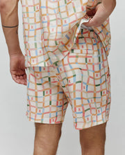 Mens Atlas Swim Trunk in cream with a multi-color plaid pattern featuring orange, blue, green, pink, and red lines, accented by small, repeated red Psycho Bunny skull logos throughout the relaxed-fit, smooth fabric.