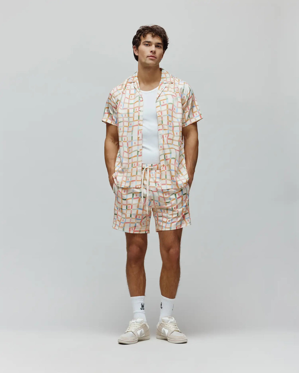 Mens Atlas Swim Trunk and matching short-sleeve camp collar shirt in a relaxed fit, featuring an off-white base with a pastel multi-color grid pattern and all-over small Psycho Bunny skull logos, plus a vertical brand text logo on the shorts.