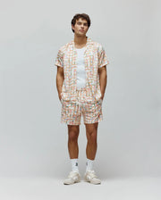 Mens Atlas Swim Trunk and matching short-sleeve camp collar shirt in a relaxed fit, featuring an off-white base with a pastel multi-color grid pattern and all-over small Psycho Bunny skull logos, plus a vertical brand text logo on the shorts.