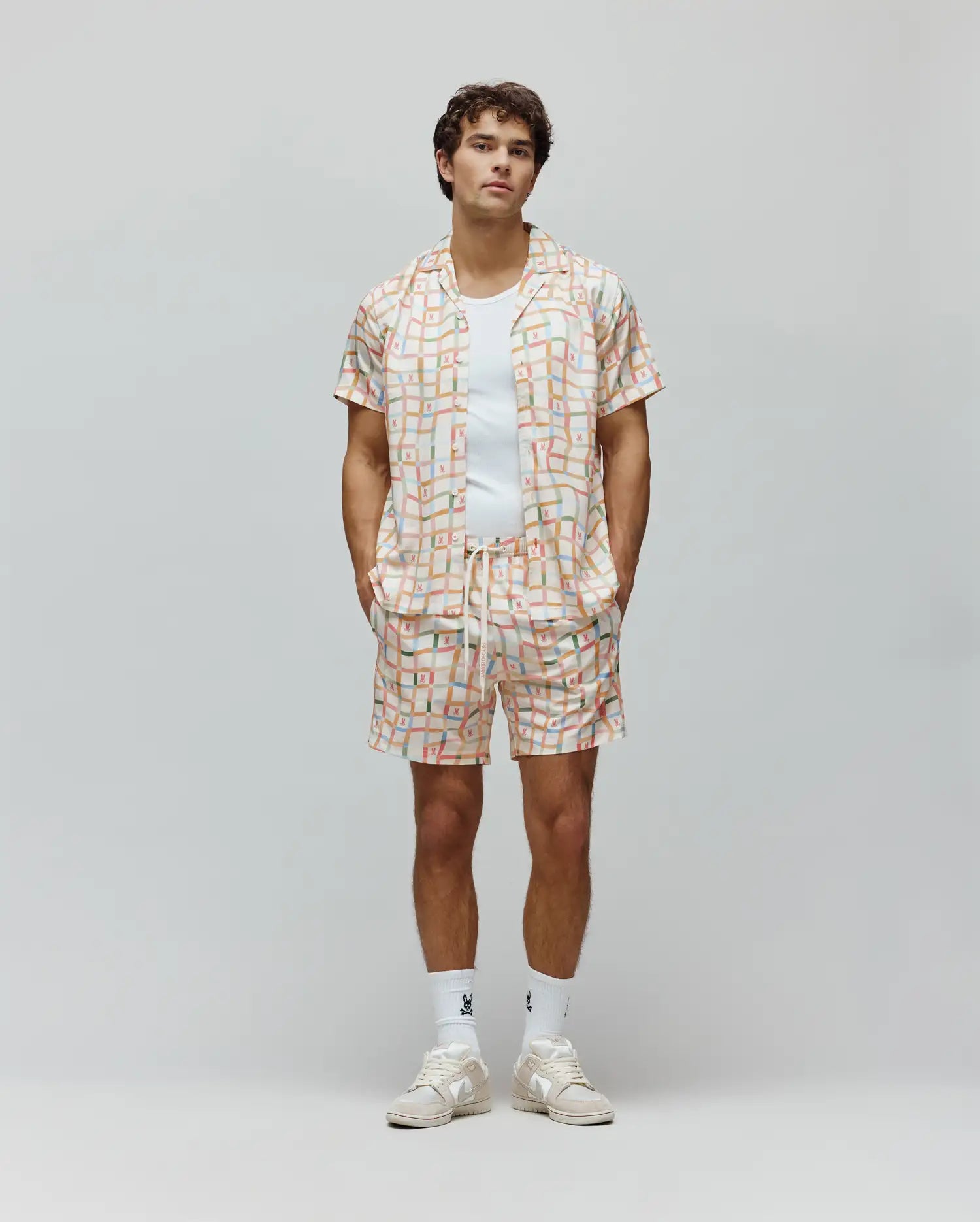 Mens Atlas Swim Trunk and matching short-sleeve camp collar shirt in a relaxed fit, featuring an off-white base with a pastel multi-color grid pattern and all-over small Psycho Bunny skull logos, plus a vertical brand text logo on the shorts.
