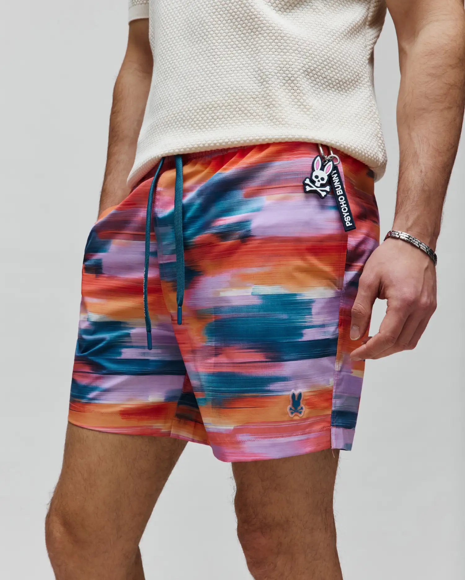 Vibrant multi-color abstract brushstroke Mens Raphael Swim Trunk with orange, pink, purple, teal hues. Features teal drawstrings, a classic fit, and a small embroidered Psycho Bunny skull logo on the left leg. A branded keychain is attached.