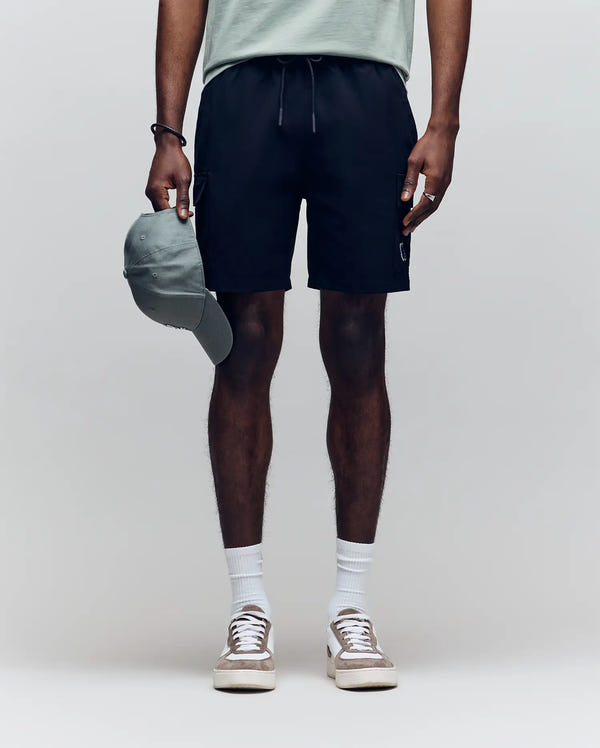 A person stands against a plain background wearing a light green T-shirt, Psycho Bunny MENS RANDY BOARD SHORT - MB1000002 with cargo pockets, white socks, and white sneakers, holding a green baseball cap. Their face is not visible.