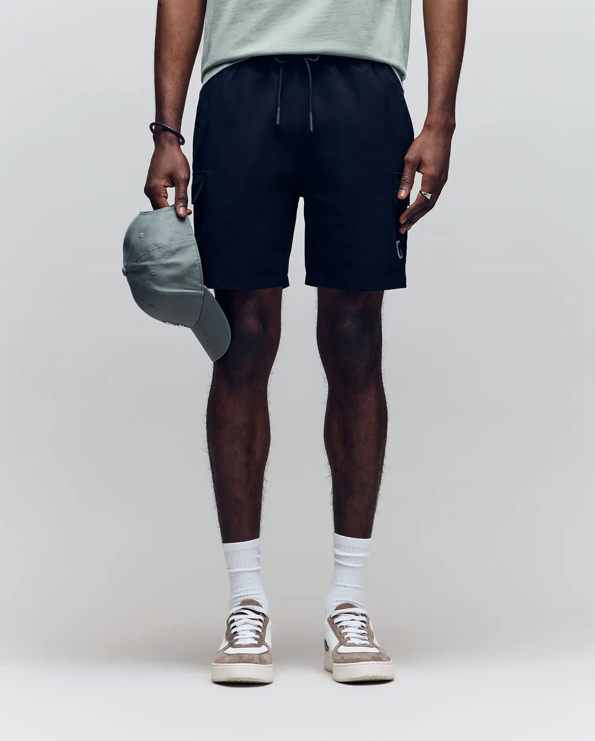 A person stands against a plain background wearing a light green T-shirt, Psycho Bunny MENS RANDY BOARD SHORT - MB1000002 with cargo pockets, white socks, and white sneakers, holding a green baseball cap. Their face is not visible.