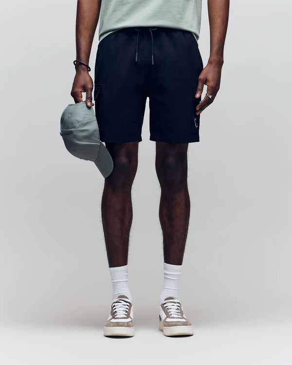 A person stands against a plain background wearing a light green T-shirt, Psycho Bunny MENS RANDY BOARD SHORT - MB1000002 with cargo pockets, white socks, and white sneakers, holding a green baseball cap. Their face is not visible.
