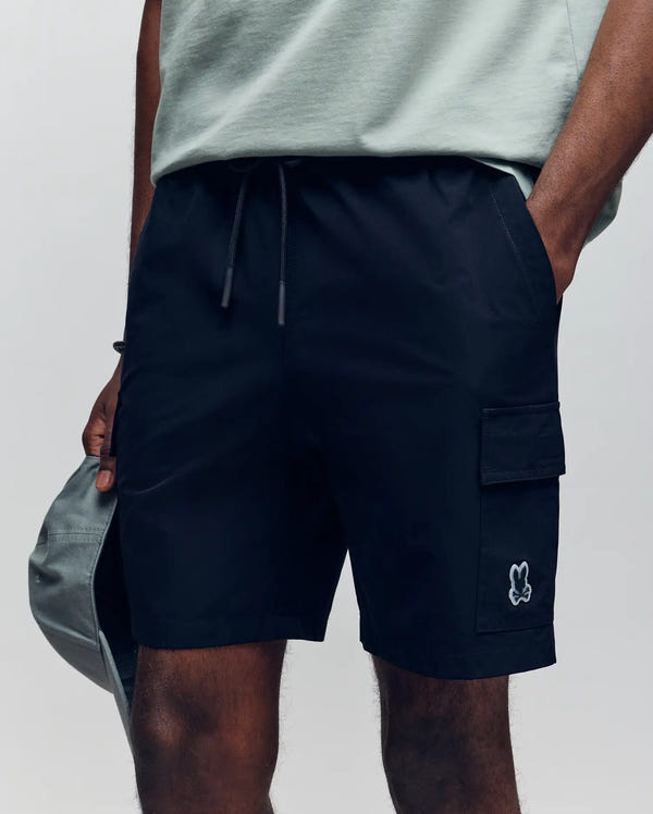 A person shown from the waist down wearing Psycho Bunny MENS RANDY BOARD SHORT - MB1000002 in black with cargo pockets and a small cartoon bunny logo, styled with a light grey T-shirt and holding a grey cap.