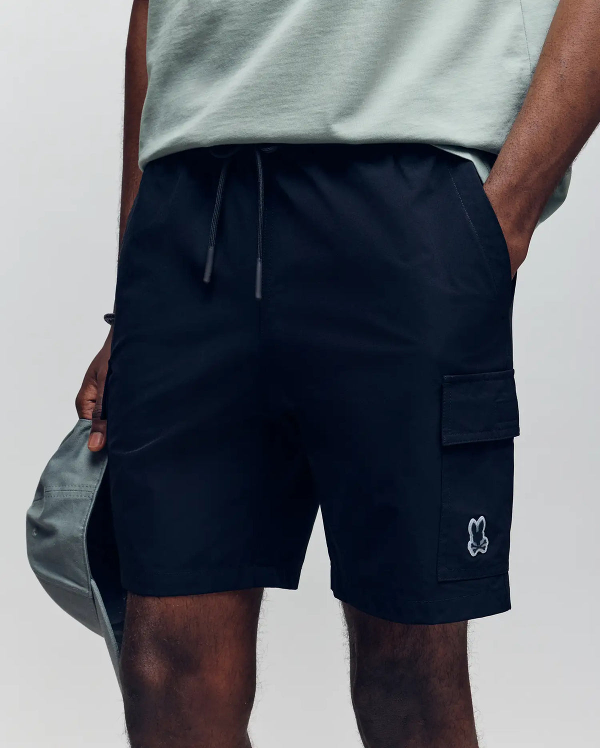 A person shown from the waist down wearing Psycho Bunny MENS RANDY BOARD SHORT - MB1000002 in black with cargo pockets and a small cartoon bunny logo, styled with a light grey T-shirt and holding a grey cap.