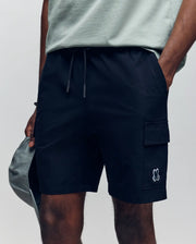 A person shown from the waist down wearing Psycho Bunny MENS RANDY BOARD SHORT - MB1000002 in black with cargo pockets and a small cartoon bunny logo, styled with a light grey T-shirt and holding a grey cap.