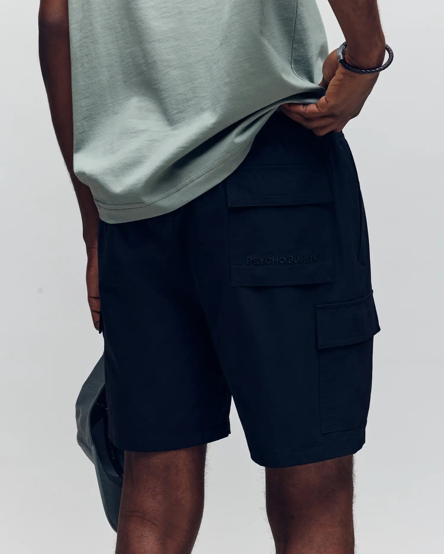A person in a light green t-shirt and Psycho Bunny MENS RANDY BOARD SHORT - MB1000002 is seen from the shoulders down, side and back view, holding a hat and showcasing the shorts’ cargo pockets.