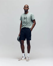 A man wearing a green T-shirt with a cartoon bunny face, Psycho Bunny MENS RANDY BOARD SHORT - MB1000002, white socks, and sneakers stands holding a cap at his side against a plain light background.