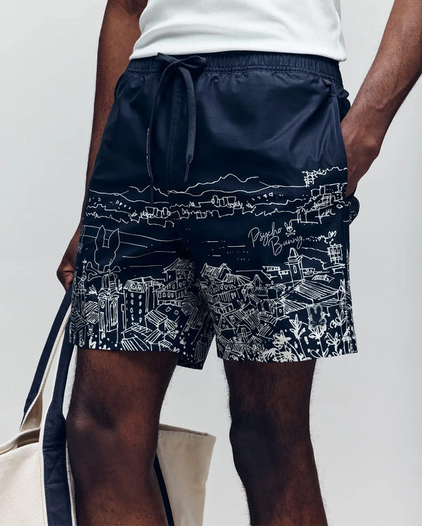 A person wears Psycho Bunny MENS NAXOS SWIM TRUNK - MB1000005 in navy, featuring a white hand-drawn cityscape print, drawstring waist, and all-day comfort, while casually holding a beige tote bag with one hand in their pocket.