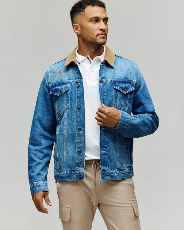 A man stands against a plain background wearing the Psycho Bunny MENS WADE DENIM TRUCKER JACKET - MO1100015, featuring a corduroy collar and subtle Bunny patch, over a white polo shirt and beige pants. He looks to the side, holding the jacket with one hand.