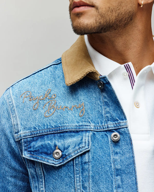 A man wears the Psycho Bunny MENS WADE DENIM TRUCKER JACKET (MO1100015) in light blue with a tan corduroy collar and embroidered logo, layered over a white collared shirt featuring purple striped placket. Only his lower face is visible.