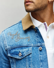 A man wears the Psycho Bunny MENS WADE DENIM TRUCKER JACKET (MO1100015) in light blue with a tan corduroy collar and embroidered logo, layered over a white collared shirt featuring purple striped placket. Only his lower face is visible.