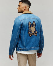 A man sports the Psycho Bunny MENS WADE DENIM TRUCKER JACKET (MO1100015) with a tan corduroy collar and a bold Bunny patch above crossed bones, styled with beige pants and a white shirt for standout street style.