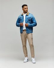 A man stands against a plain background wearing the Psycho Bunny MENS WADE DENIM TRUCKER JACKET - MO1100015 with a tan corduroy collar, paired with a white shirt, beige cargo pants, and white sneakers. He looks to the side, holding his jacket’s front.