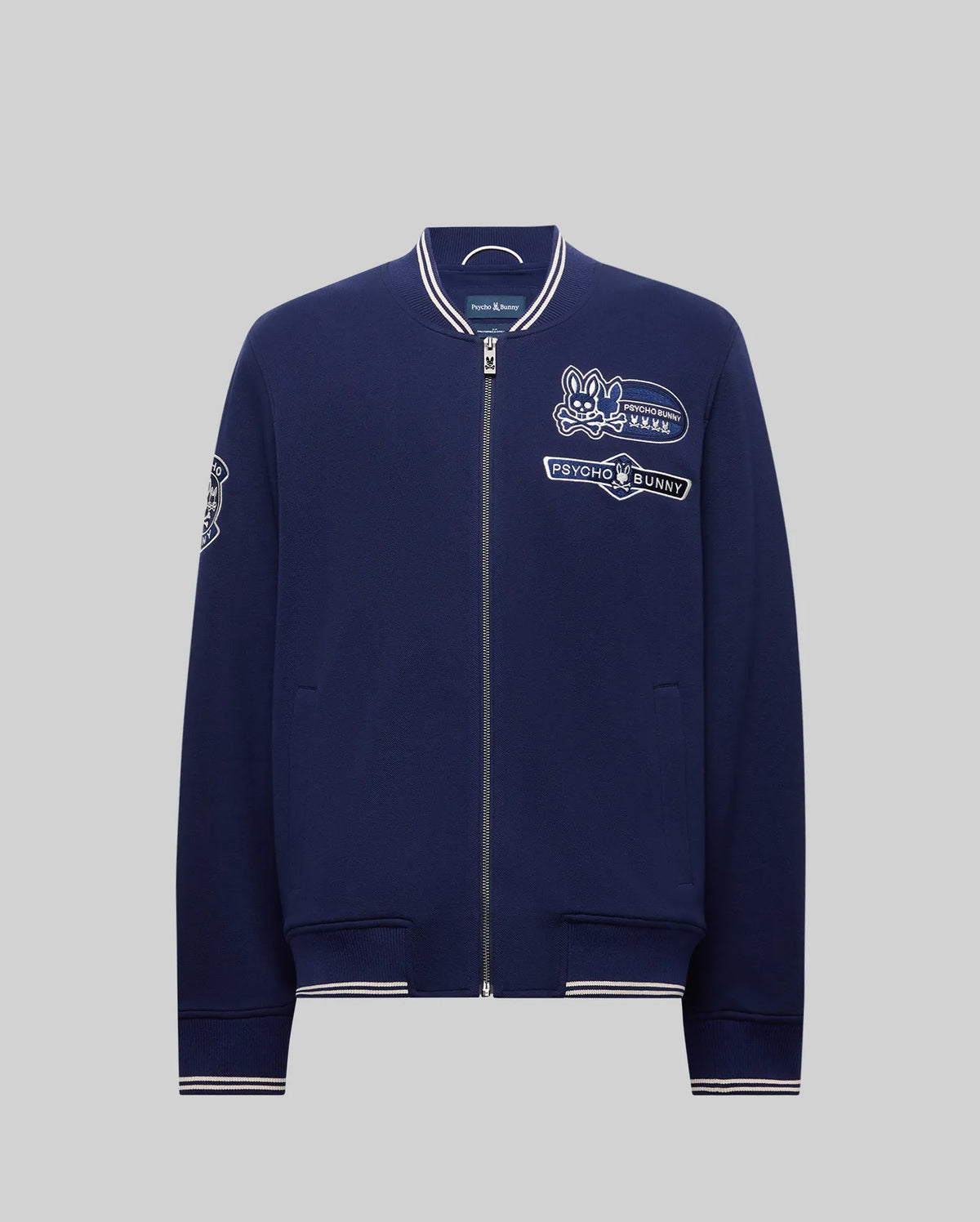 The Psycho Bunny MENS DESMOND PIQUE KNIT BOMBER - MO1100018 is a navy bomber with white trim, varsity-style details, and embroidered patches including a bunny logo, "Psycho Bunny" text on the chest, and a sleeve patch.