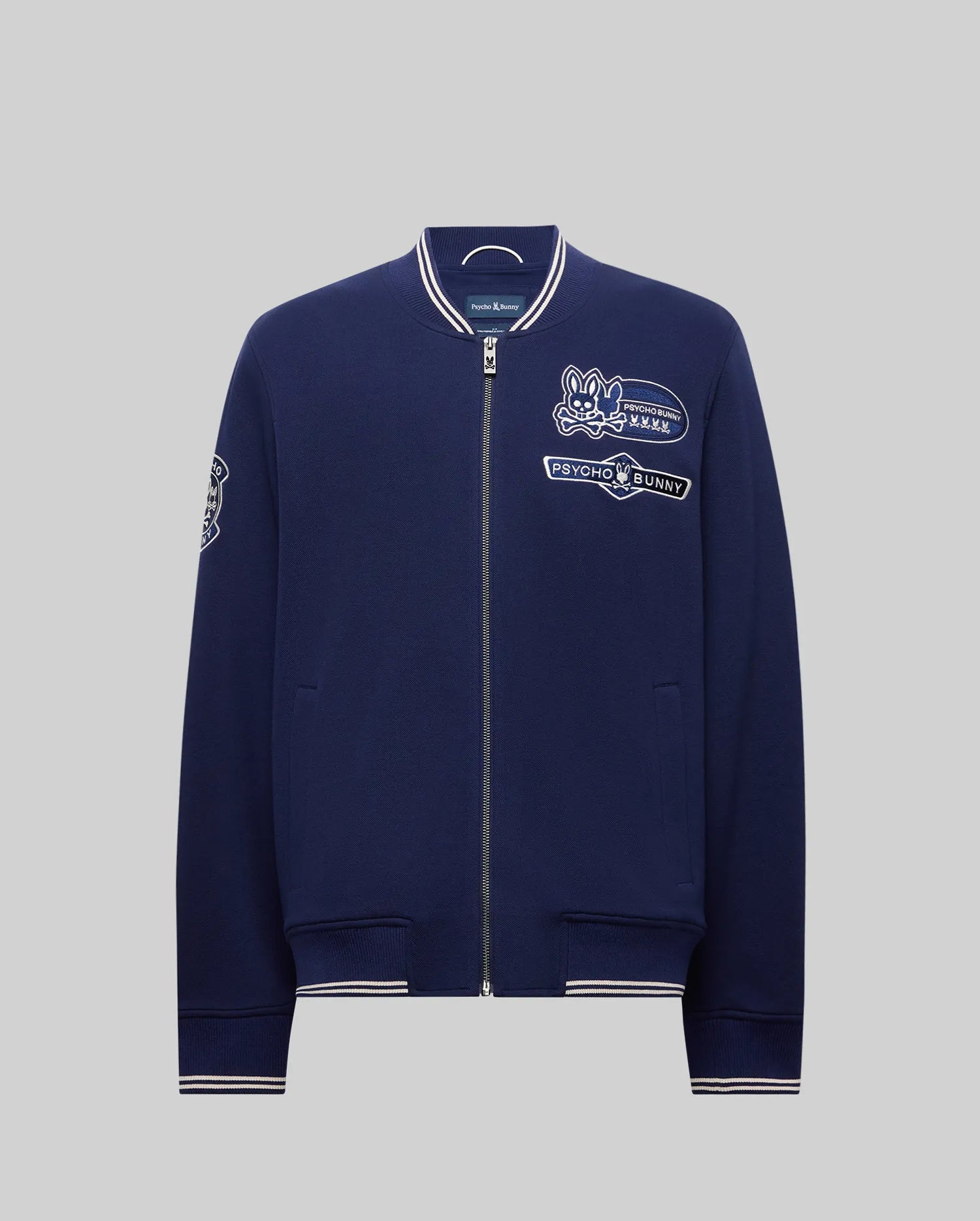 The Psycho Bunny MENS DESMOND PIQUE KNIT BOMBER - MO1100018 is a navy bomber with white trim, varsity-style details, and embroidered patches including a bunny logo, "Psycho Bunny" text on the chest, and a sleeve patch.