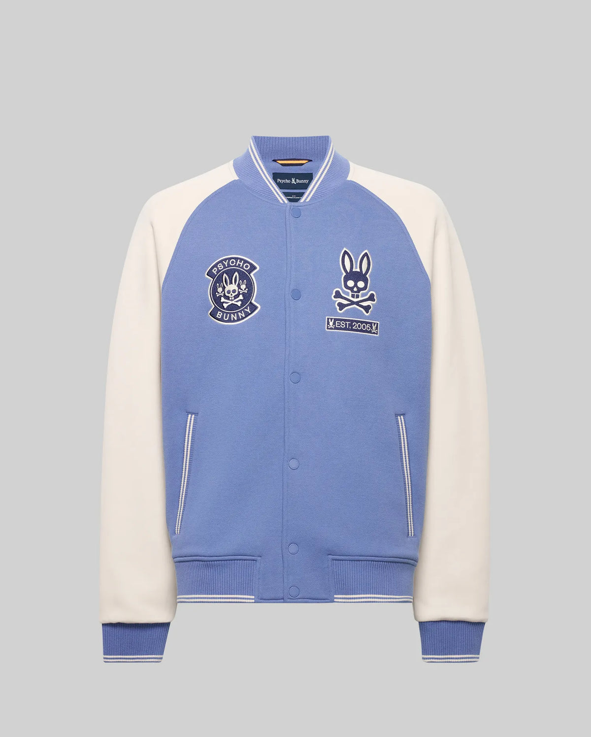 The Psycho Bunny MENS RYDER KNIT BOMBER (MO1100034) features light blue body, white sleeves, striped blue cuffs and collar, bunny patches, and an embroidered chest logo, shown against a plain grey background.