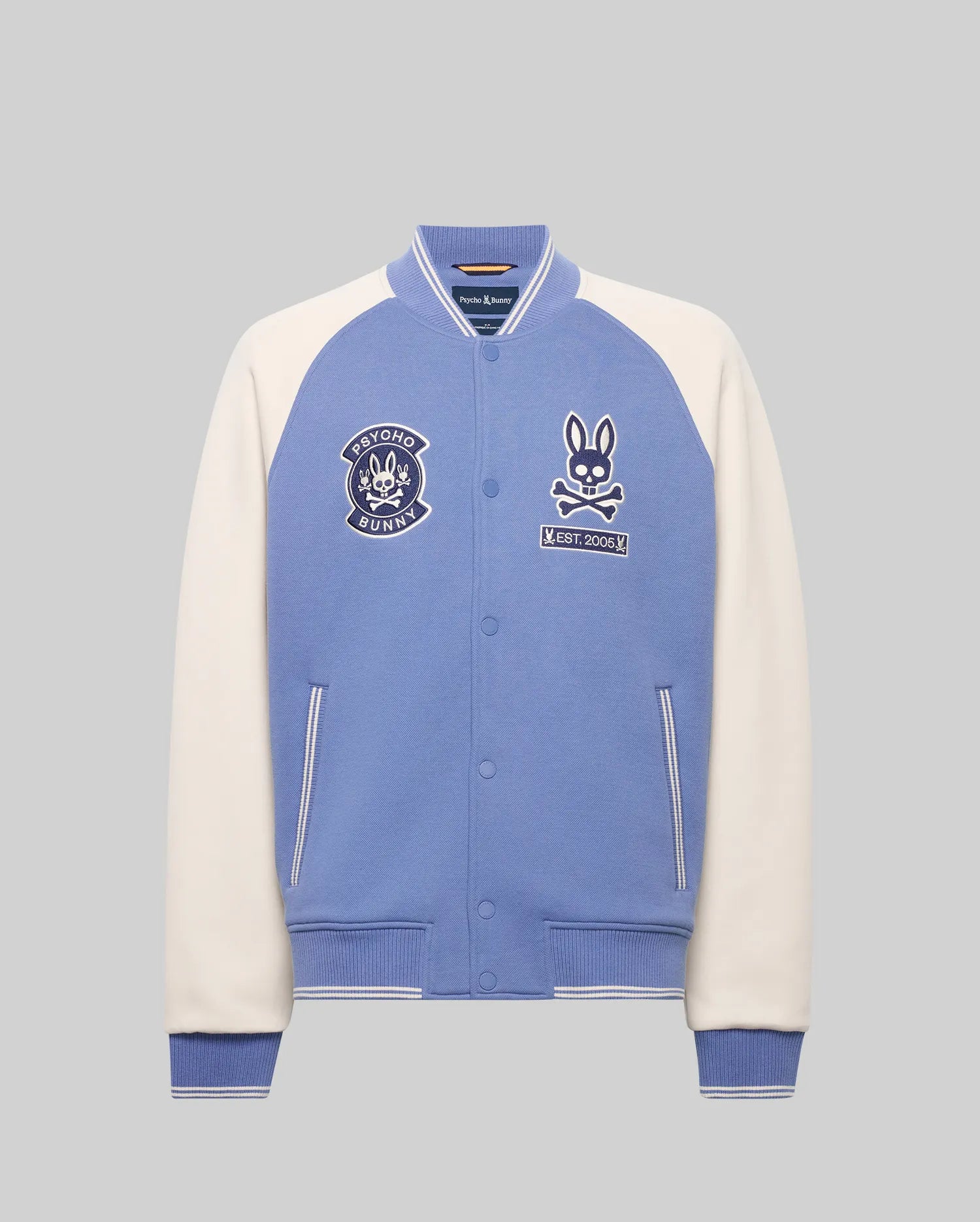The Psycho Bunny MENS RYDER KNIT BOMBER (MO1100034) features light blue body, white sleeves, striped blue cuffs and collar, bunny patches, and an embroidered chest logo, shown against a plain grey background.