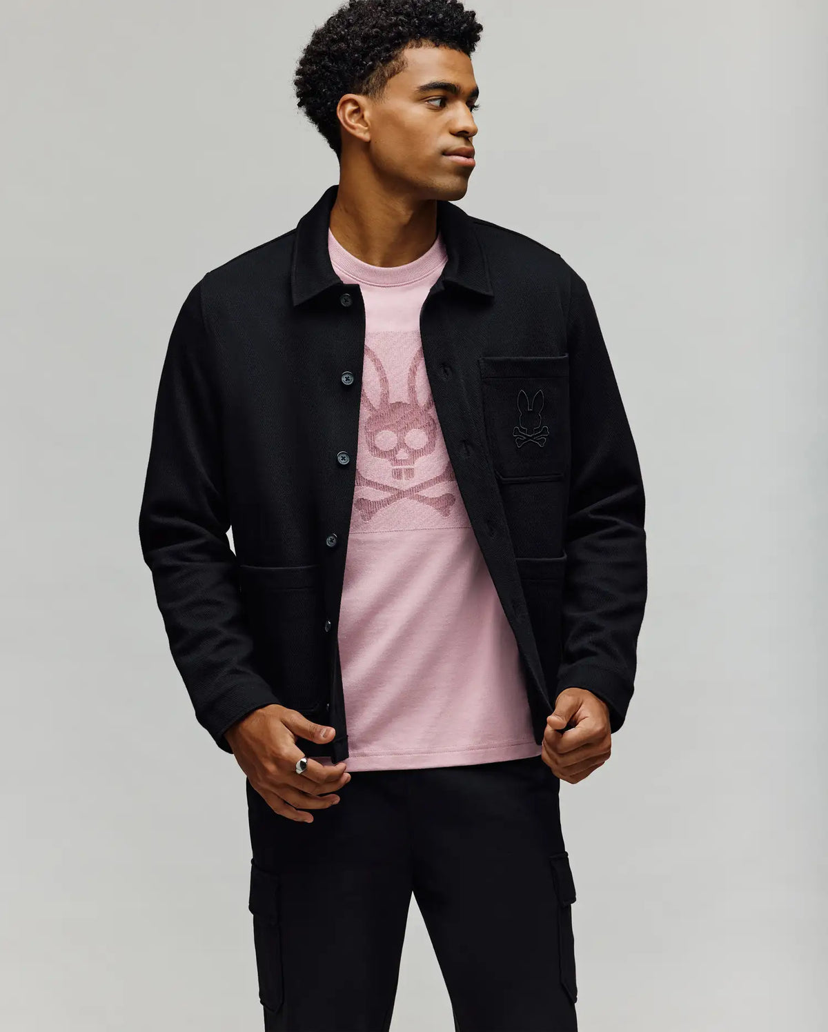 A man wears the Psycho Bunny MENS AVEN KNIT SHACKET (MO1100035) in black over a light pink bunny graphic tee, standing against a plain backdrop with one hand opening his jacket—an effortless, comfortable look.