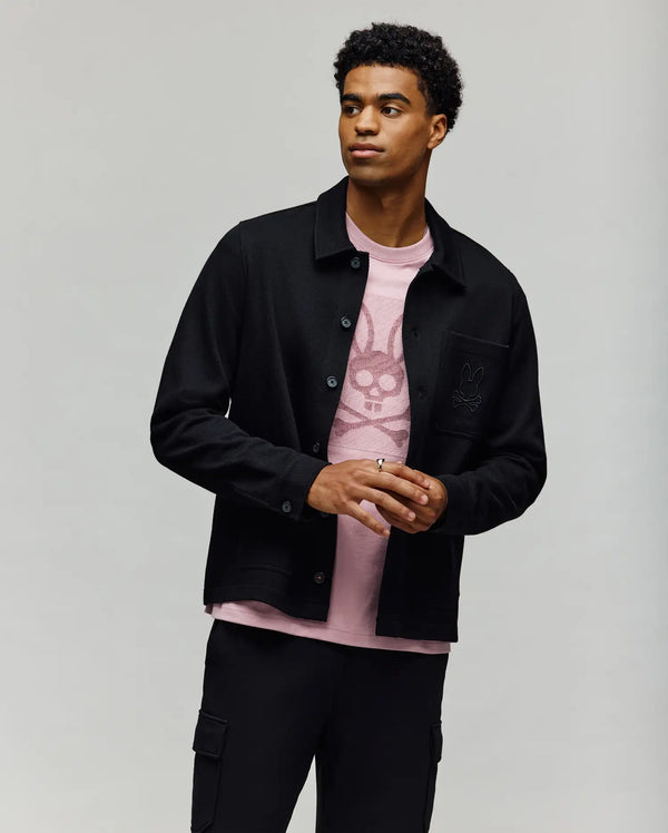 A man with short curly hair wears the Psycho Bunny MENS AVEN KNIT SHACKET (MO1100035) over a light pink bunny graphic tee and black cargo pants, standing against a plain light background.