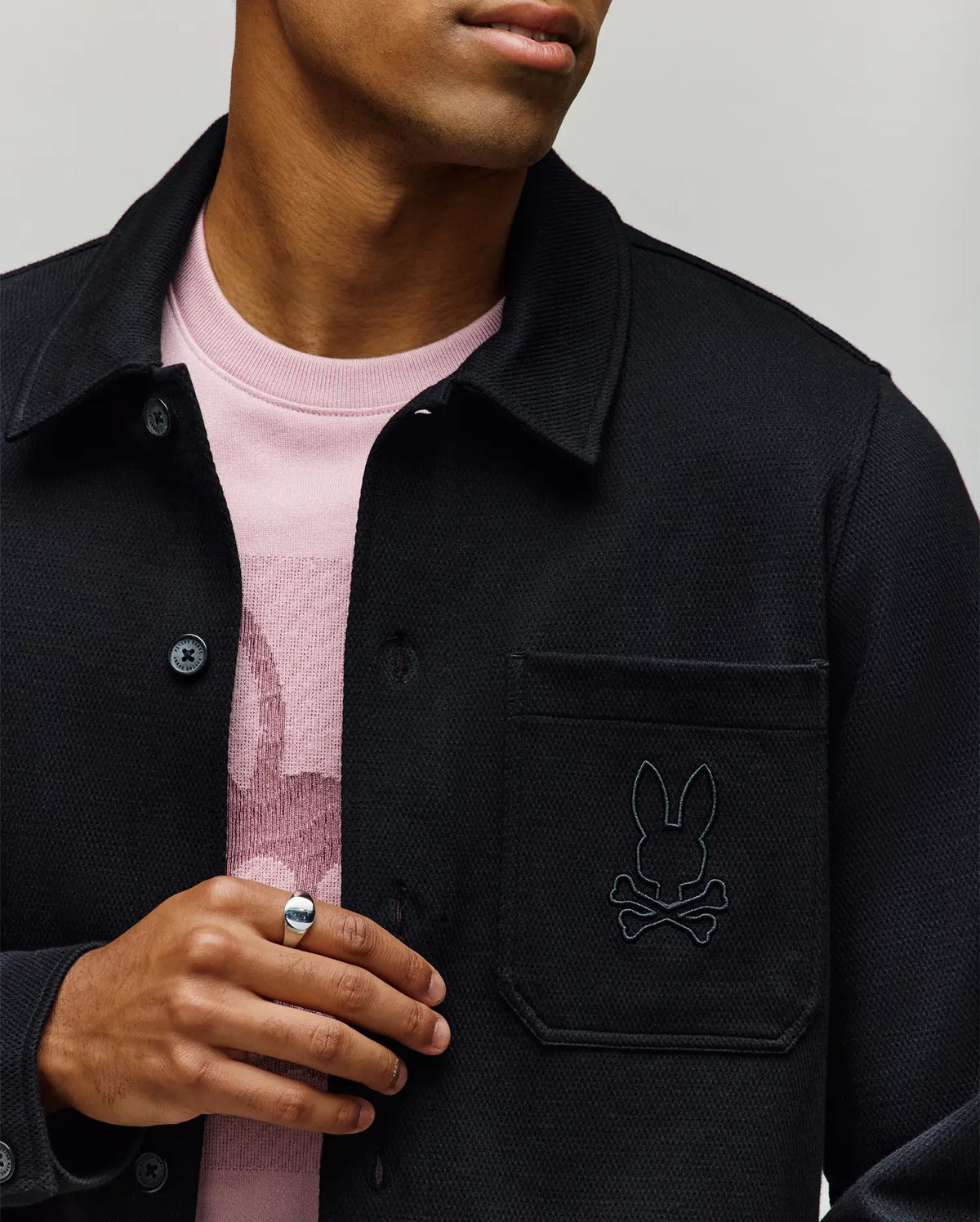 A person wears the Psycho Bunny MENS AVEN KNIT SHACKET (MO1100035) in black, featuring the bunny and crossbones logo on the pocket, layered over a light pink shirt. The look is styled with a silver ring and shown from shoulders to chest.