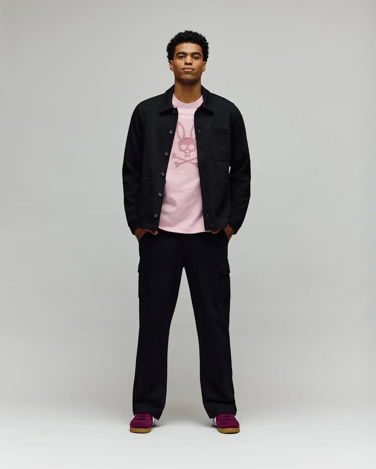 A man faces forward in a Psycho Bunny MENS AVEN KNIT SHACKET (MO1100035), styled over a pink graphic tee, black cargo pants, and dark pink sneakers, effortlessly displaying transitional comfort against a plain light gray background.