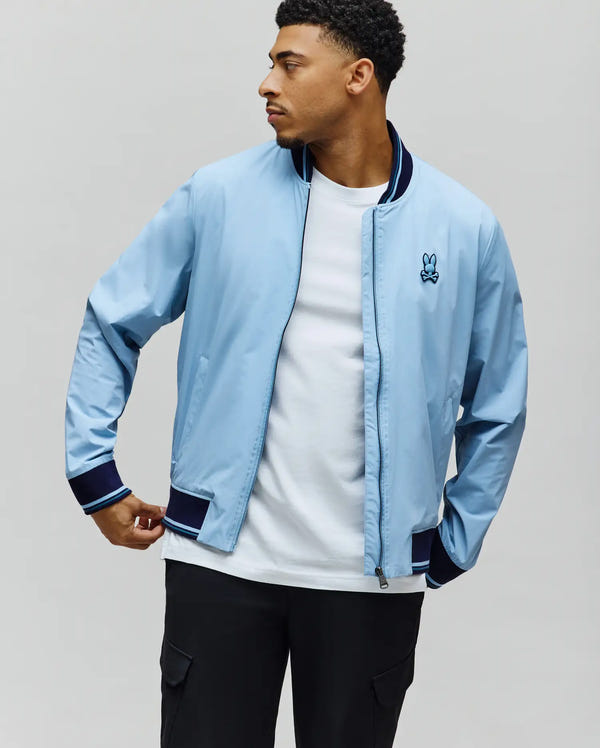 MENS ALDRIN BOMBER in sky blue, a classic fit jacket with full zip, navy ribbed collar, cuffs, and hem featuring light blue stripes, and an embroidered Psycho Bunny skull logo on the left chest.