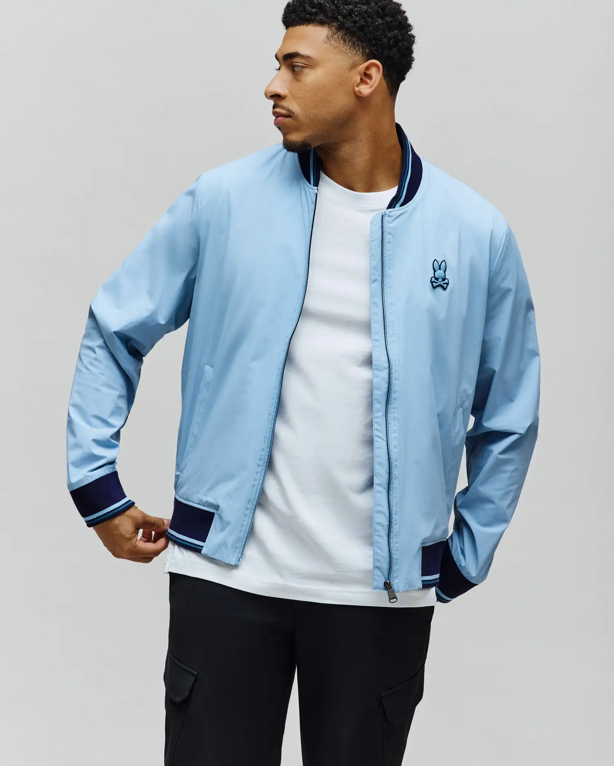 MENS ALDRIN BOMBER in sky blue, a classic fit jacket with full zip, navy ribbed collar, cuffs, and hem featuring light blue stripes, and an embroidered Psycho Bunny skull logo on the left chest.