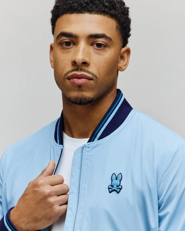 Mens Aldrin Bomber, light blue with navy and light blue striped ribbed collar and cuffs, full zip, featuring an embroidered Psycho Bunny skull logo on the left chest.