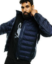 Mens Emerik Hooded Puffer Jacket in dark navy, with horizontal quilting and full zip. Layered over a black jersey t-shirt featuring a large grey Psycho Bunny skull logo with subtle pattern on the chest. (3)