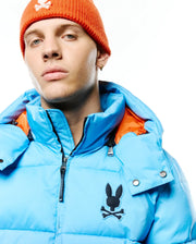 Men's Solan Puffer Jacket in bright blue with a contrasting orange hood lining, featuring a large black Psycho Bunny skull logo on the left chest and a full zip closure. (4)