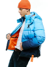 Men's Solan Puffer Jacket, an ombre light to dark blue padded jacket with a navy bottom, features a large black embroidered Psycho Bunny skull logo on the left chest, bright orange lining, and a branded orange tag. (5)