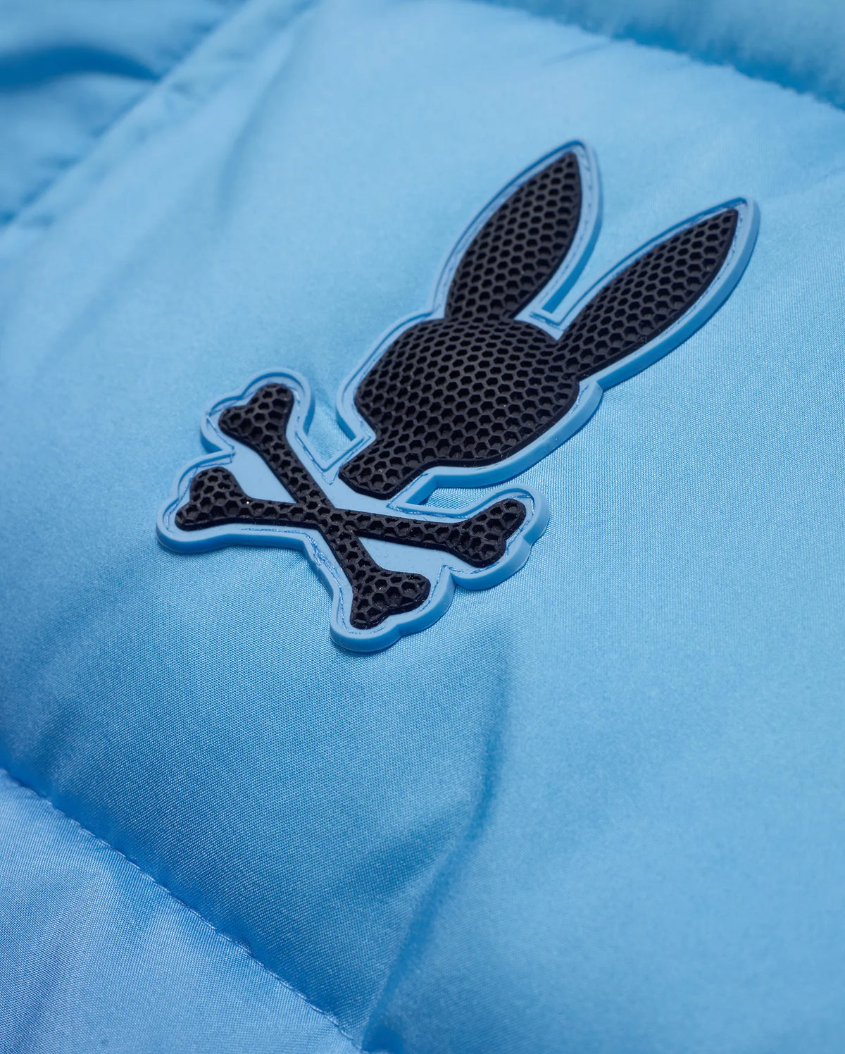 Men's Solan Puffer Jacket in sky blue, featuring a smooth, padded nylon texture and a rubberized dark grey Psycho Bunny skull logo with a light blue outline on the chest. (8)