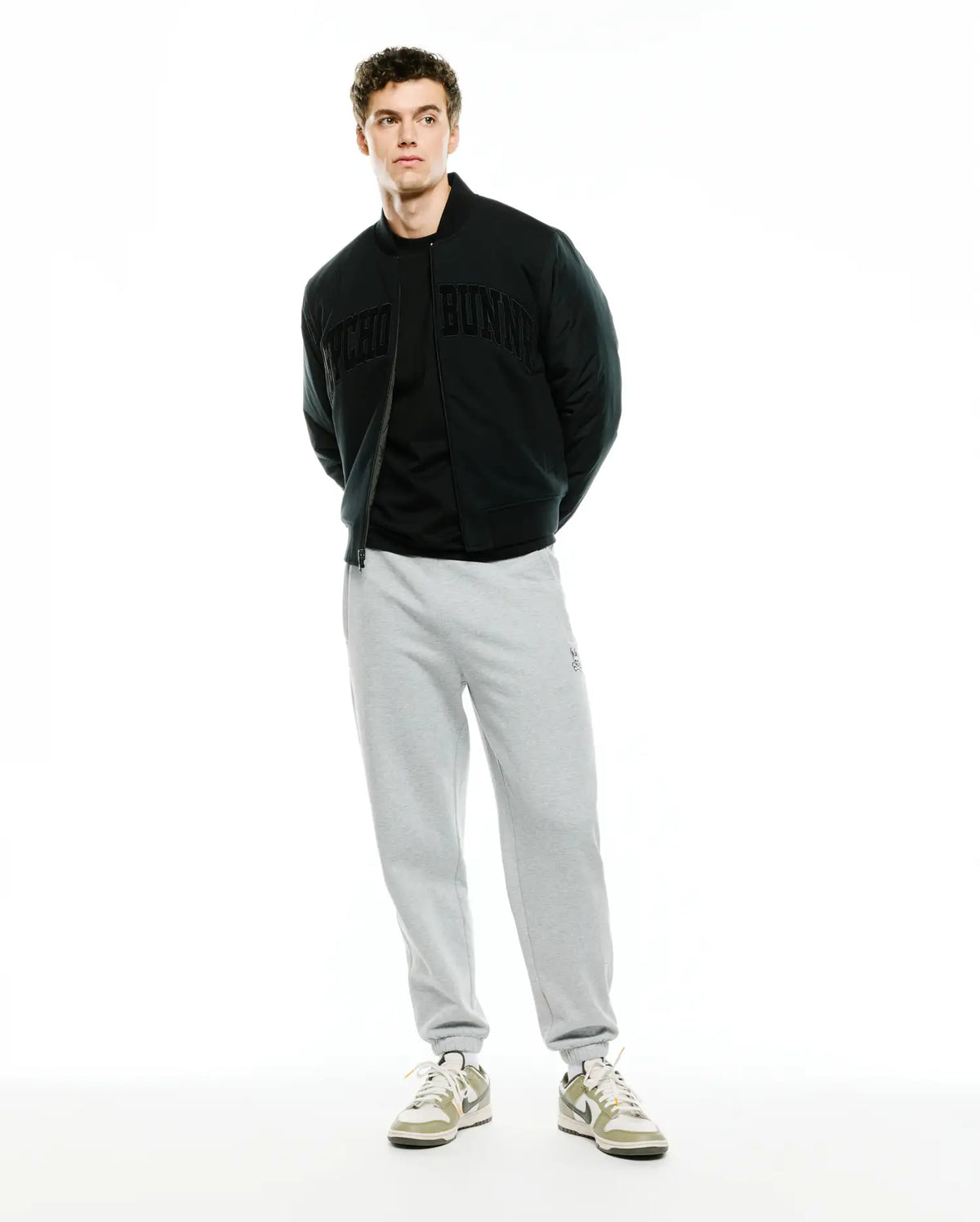 Mens Joaquin Mix Media Reversible Bomber in black, featuring large tonal Psycho Bunny lettering across the chest, paired with light grey relaxed fit french terry sweatpants with a small embroidered bunny skull logo on the left thigh. (5)