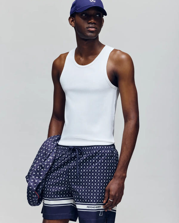 A man in a Psycho Bunny MENS 2 PACK TANK TOP - MO1400001, navy cap, and patterned swim shorts holds a matching jacket while standing against a plain grey background.