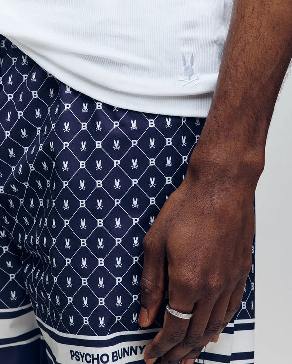 A close-up shows a person in a white shirt and navy blue Psycho Bunny shorts, with a silver ring on their hand. The look features the brand’s MENS 2 PACK TANK TOP - MO1400001.