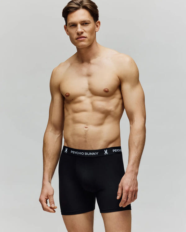 A fit, shirtless man wears Psycho Bunny MENS 3 PACK BOXER BRIEF - MO1400006 in black with a logo waistband, made from a cotton-modal blend, posing against a plain background.